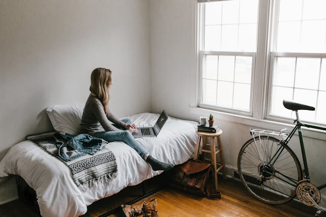 freelancer in a room with a laptop on a bed facing the window