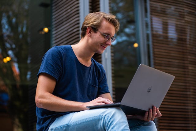 freelancer looking at his laptop while sitting with building on the background